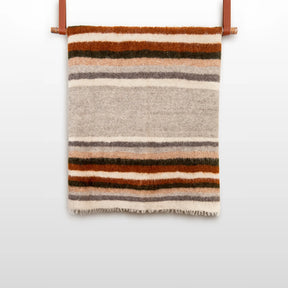 The Mudry (Wise) Wool Statement Bed Blanket, made of organic Carpathian wool and featuring brown, gray, and cream stripes, is folded and hangs on a leather strap against a white wall.