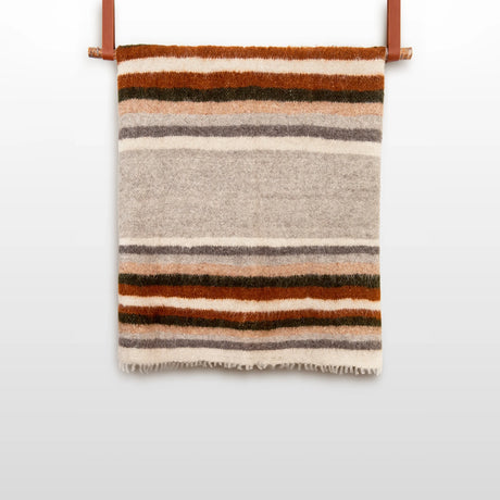 The Mudry (Wise) Wool Statement Bed Blanket, made of organic Carpathian wool and featuring brown, gray, and cream stripes, is folded and hangs on a leather strap against a white wall.