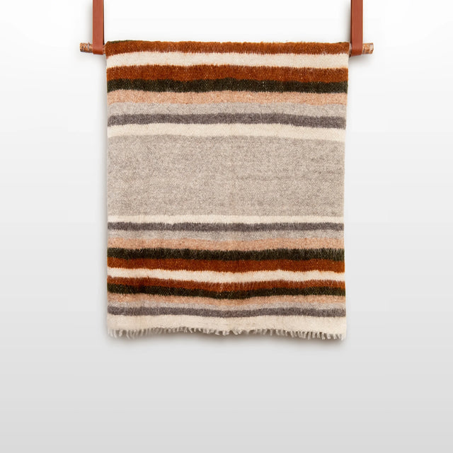 The Mudry (Wise) Wool Statement Bed Blanket, made of organic Carpathian wool and featuring brown, gray, and cream stripes, is folded and hangs on a leather strap against a white wall.
