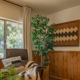 Cozy corner with an armchair, pillows, a potted plant, window, and the Dobre (Good) Wool Framed Wall Art featuring a geometric pattern above wood paneling.
