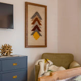 A cozy corner with an olive armchair, blanket, blue cabinet, and a Strilka (Arrow) Wool Framed Wall Art featuring geometric designs above.