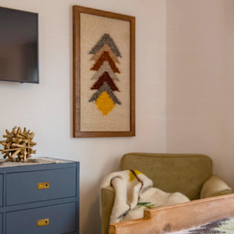 A cozy corner with an olive armchair, blanket, blue cabinet, and a Strilka (Arrow) Wool Framed Wall Art featuring geometric designs above.