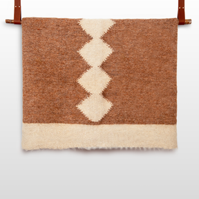 The Druh (Friend) Wool Statement Bed Blanket, in brown and cream undyed wool with a zigzag diamond pattern, hangs from two brown leather straps.