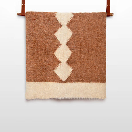 The Druh (Friend) Wool Statement Bed Blanket, woven in brown and white from organic cruelty-free wool with a central diamond pattern, hangs by two leather straps against a white wall.