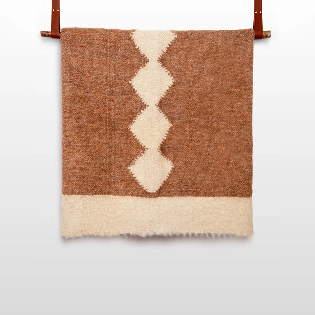 The Druh (Friend) Wool Statement Bed Blanket, woven in brown and white from organic cruelty-free wool with a central diamond pattern, hangs by two leather straps against a white wall.