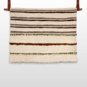 The Faino (Fine) Wool Statement Bed Blanket features horizontal brown, green, and beige stripes and leather straps—handcrafted wool reimagined as striking wall art.