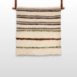 The Faino (Fine) Wool Statement Bed Blanket features horizontal stripes in brown, green, and cream, crafted to resemble a handwoven textile and accented with two leather straps for hanging.