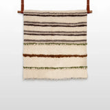 The Faino (Fine) Wool Statement Bed Blanket features horizontal stripes in brown, green, and cream, crafted to resemble a handwoven textile and accented with two leather straps for hanging.