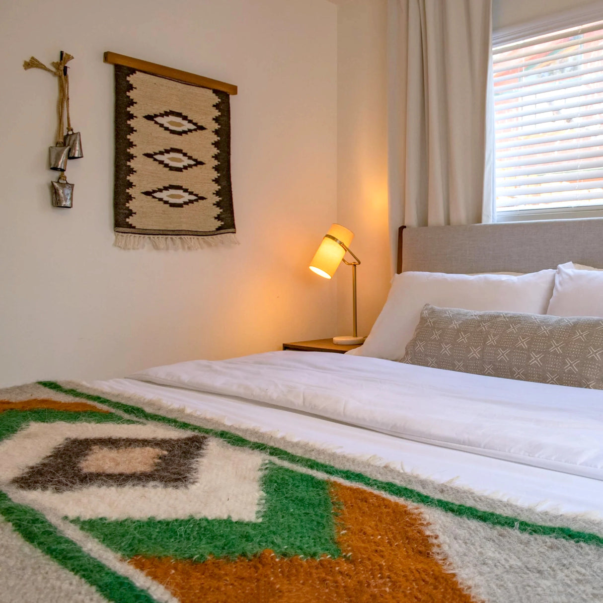 Cozy bedroom featuring the Heredij (Heritage) Wool Rug as a wall hanging, a patterned blanket, wall art, bedside lamp, and a window with blinds letting in natural light.