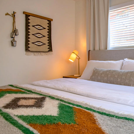 Cozy bedroom featuring the Heredij (Heritage) Wool Rug as a wall hanging, a patterned blanket, wall art, bedside lamp, and a window with blinds letting in natural light.