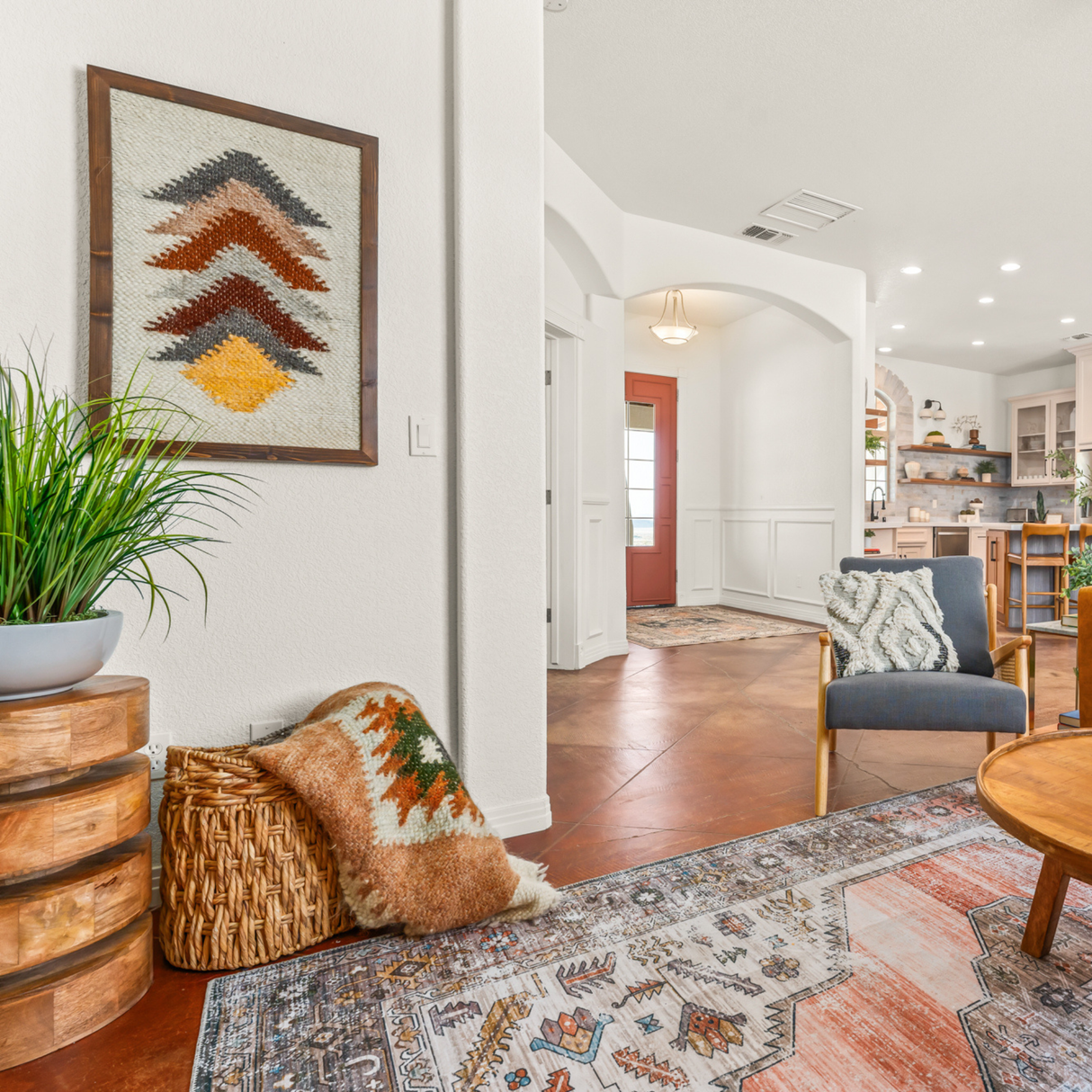 Cozy living room with patterned rug, Strilka (Arrow) Wool Framed Wall Art, potted plant, comfy pillows, and a view into a bright kitchen.