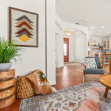 Cozy living room with patterned rug, Strilka (Arrow) Wool Framed Wall Art, potted plant, comfy pillows, and a view into a bright kitchen.