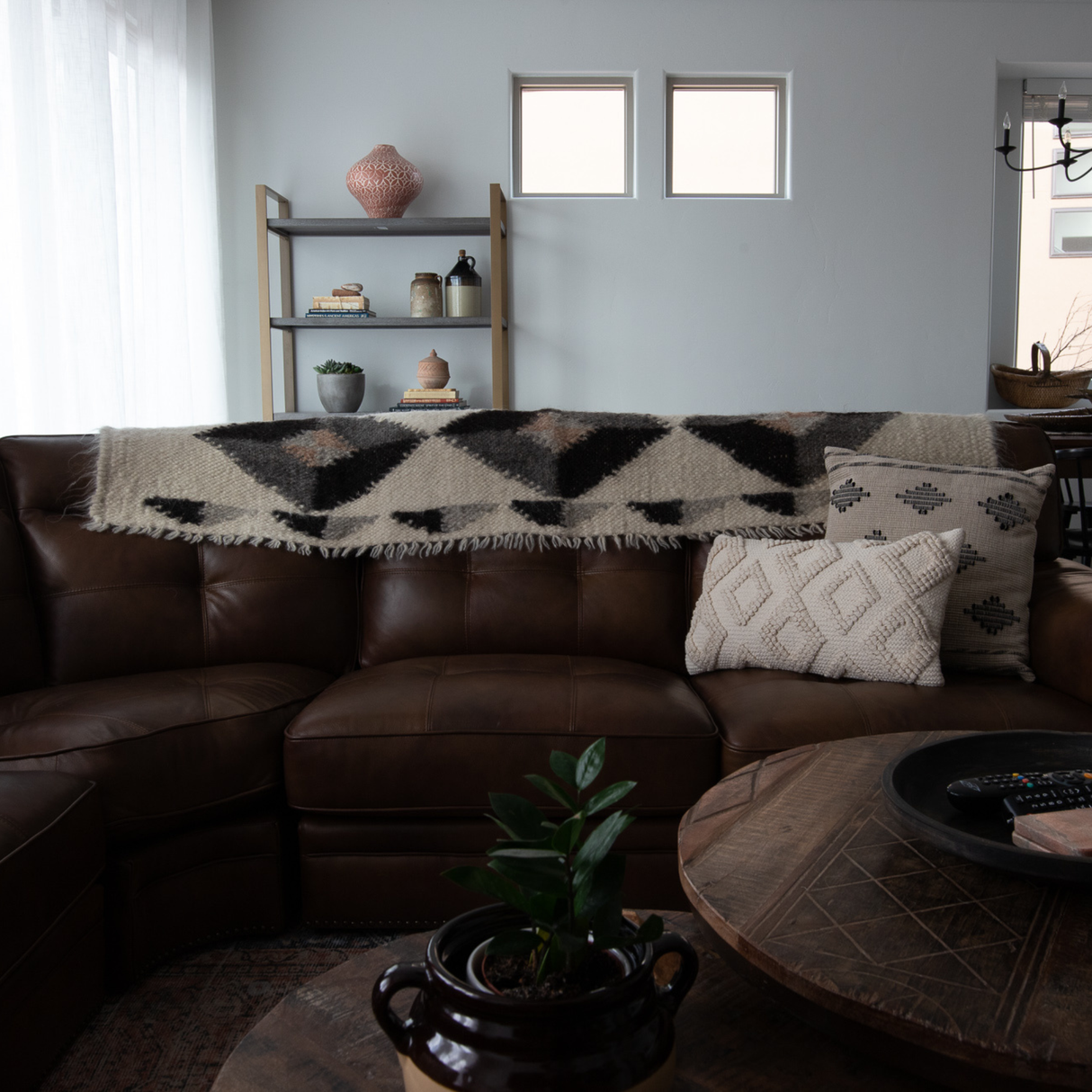 Brown leather sectional with patterned throws and pillows, an Iskara (Spark) Wool Bed/Couch Runner from the Natural Collection, wooden coffee table, shelves, and a potted plant with undyed wool accents.