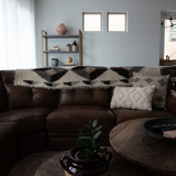 Brown leather sectional with patterned throws and pillows, an Iskara (Spark) Wool Bed/Couch Runner from the Natural Collection, wooden coffee table, shelves, and a potted plant with undyed wool accents.