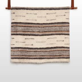 The Gratsia (Grace) Wool Statement Bed Blanket in cream and brown, with horizontal stripes and textured patterns, hangs on a wooden rod—a cozy, handcrafted accent for any space.