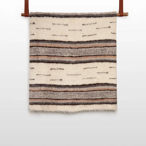 The Gratsia (Grace) Wool Statement Bed Blanket, crafted from organic wool in cream with brown and gray stripes, hangs elegantly on a wooden rod.