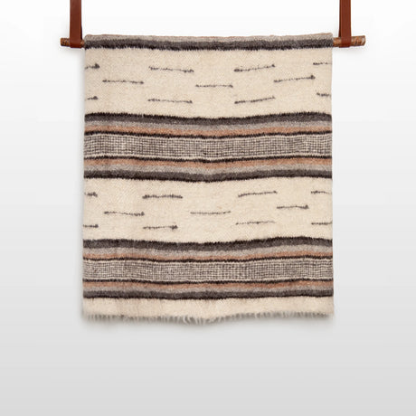 The Gratsia (Grace) Wool Statement Bed Blanket, crafted from organic wool in cream with brown and gray stripes, hangs elegantly on a wooden rod.