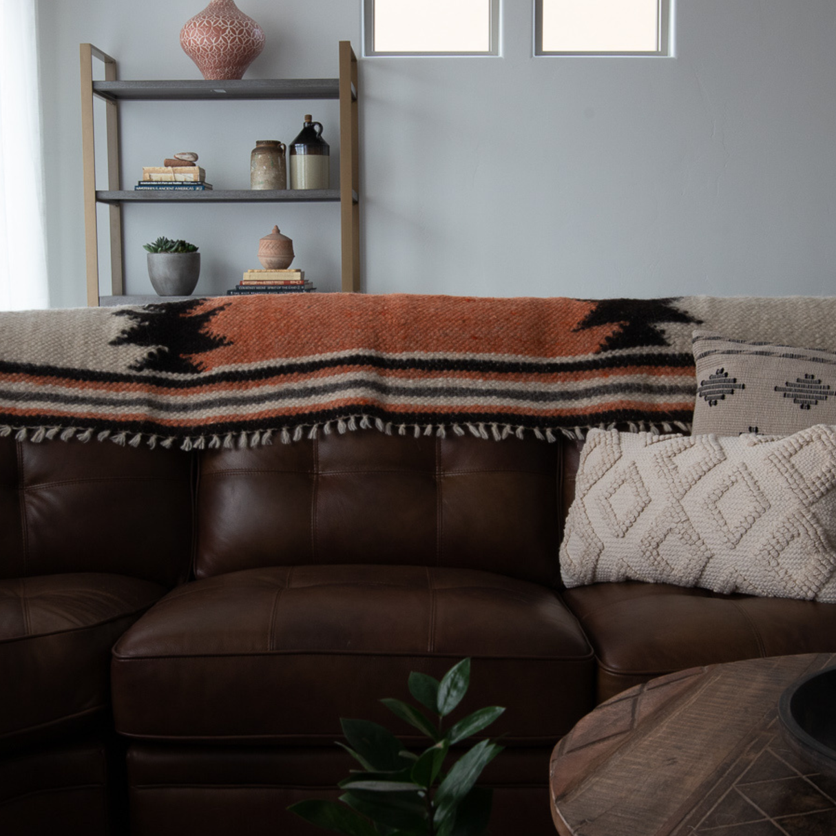 A brown leather sofa with patterned pillows, a handcrafted Opora (Pillar) Wool Bed/Couch Runner, a shelf with decor, and a plant in front.