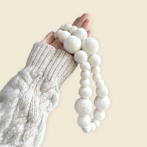 A hand in a cream knit sleeve holds the 8' Hand-felted Natural Cream Organic Wool Garland, featuring white organic New Zealand wool balls in varying sizes.