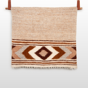 The Tkach (Weaver) Wool Statement Bed Blanket features brown, beige, and white geometric diamond patterns and is shown hanging on a wooden rod.