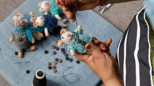 Hands sewing a button onto a small felt mouse toy, with other similar toys and buttons nearby.