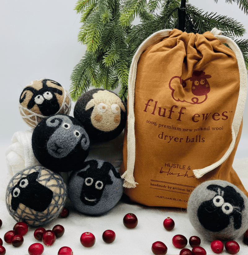 Six sheep-themed wool dryer balls, a brown drawstring bag, pine branches, and cranberries on a white surface.