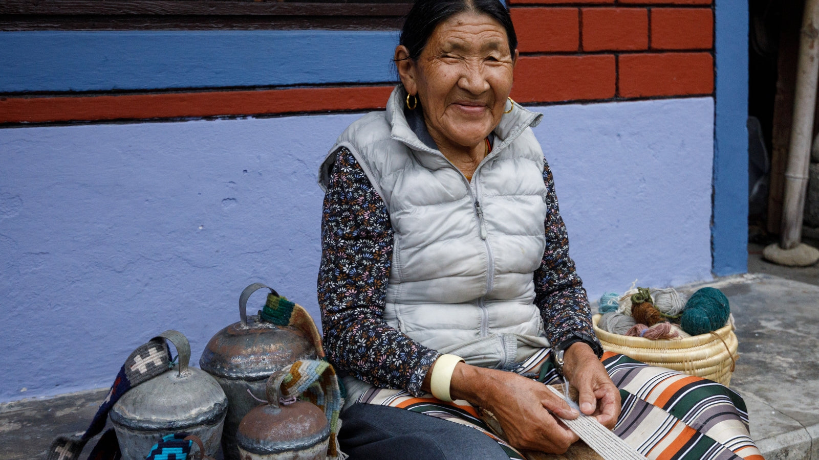 An elderly woman smiles while weaving, seated by colorful yarn and containers against a blue and red wall.