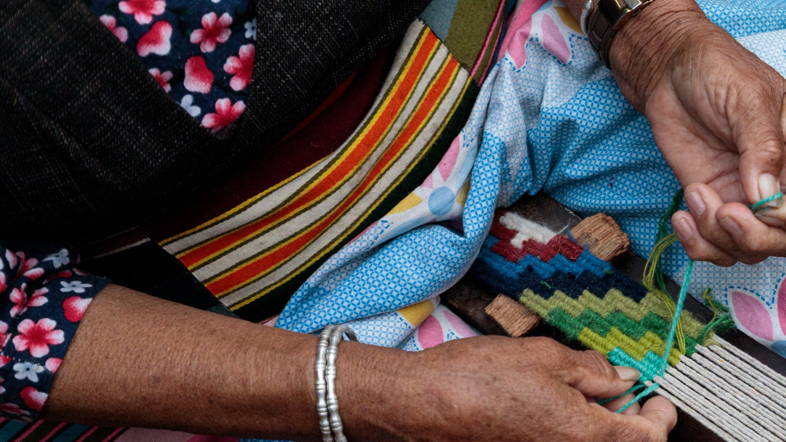 Close-up of an older person weaving colorful fabric by hand, wearing bright patterned clothing and jewelry.