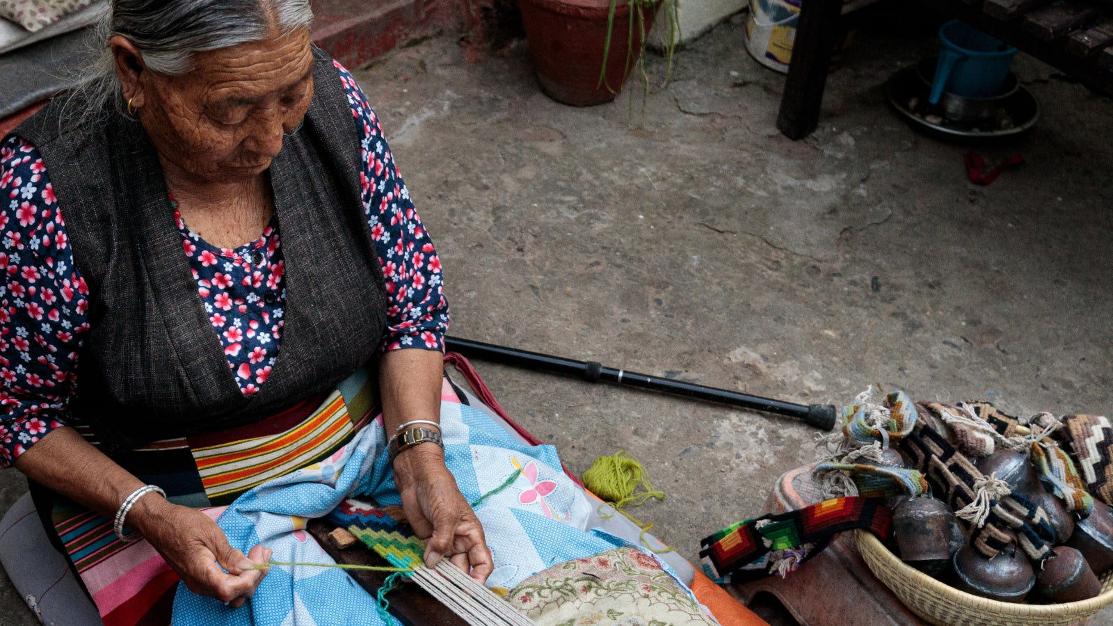 Elderly woman sitting outdoors, weaving with colorful yarn, surrounded by textiles and household items.