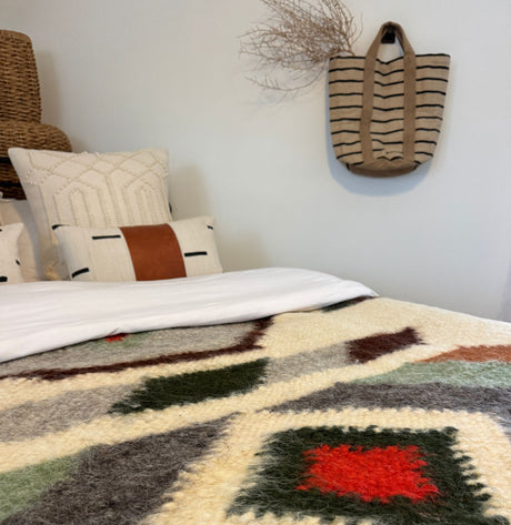 A cozy bed features the Diamant (Diamond) Wool Statement Bed Blanket made from organic, cruelty-free wool; patterned pillows and a striped tote bag complete the look.