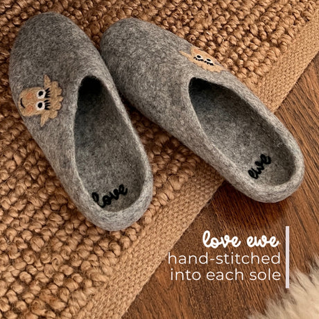 Love Ewe Hand-Felted Organic Wool Slippers for Men (Blush-LG), featuring sheep designs and "love ewe" hand-stitched inside, offer fair trade comfort and charm—perfect for relaxing at home. Placed on a woven mat.