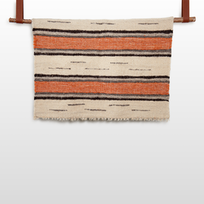 The Chudovo (Well) Wool Statement Throw / Baby Blanket, handcrafted from Carpathian sheep wool with orange and black stripes, hangs on a wooden rod against a white background.