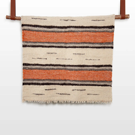 The Chudovo (Well) Wool Statement Throw / Baby Blanket, featuring orange and black horizontal stripes, hangs on a rod against a white background.