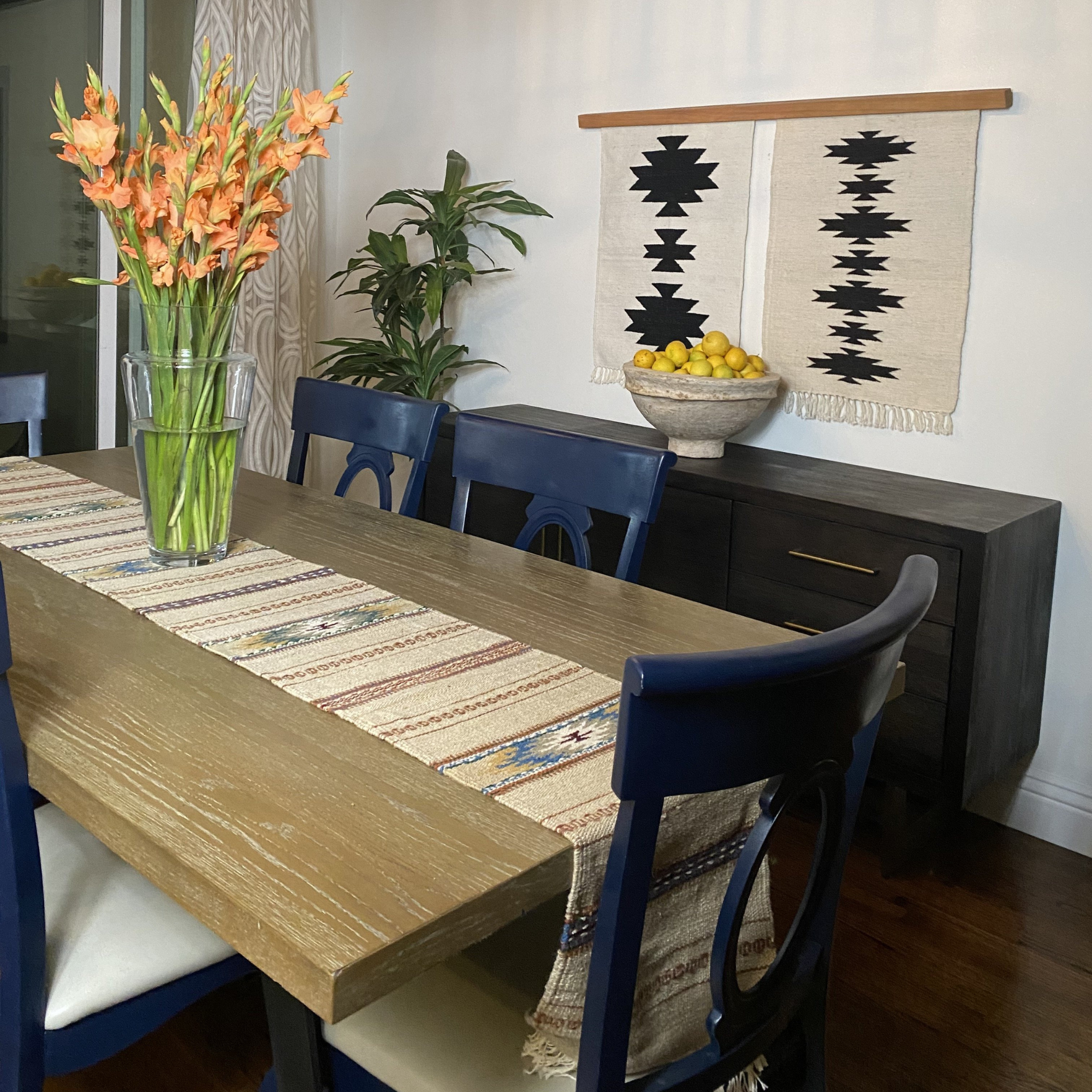Dining room with a wooden table, blue chairs, orange flowers, and the Chysto Wool Rug/Wall Hanging Duo with Wood Rug Hanger above a dark sideboard for a stylish, eco-friendly touch.