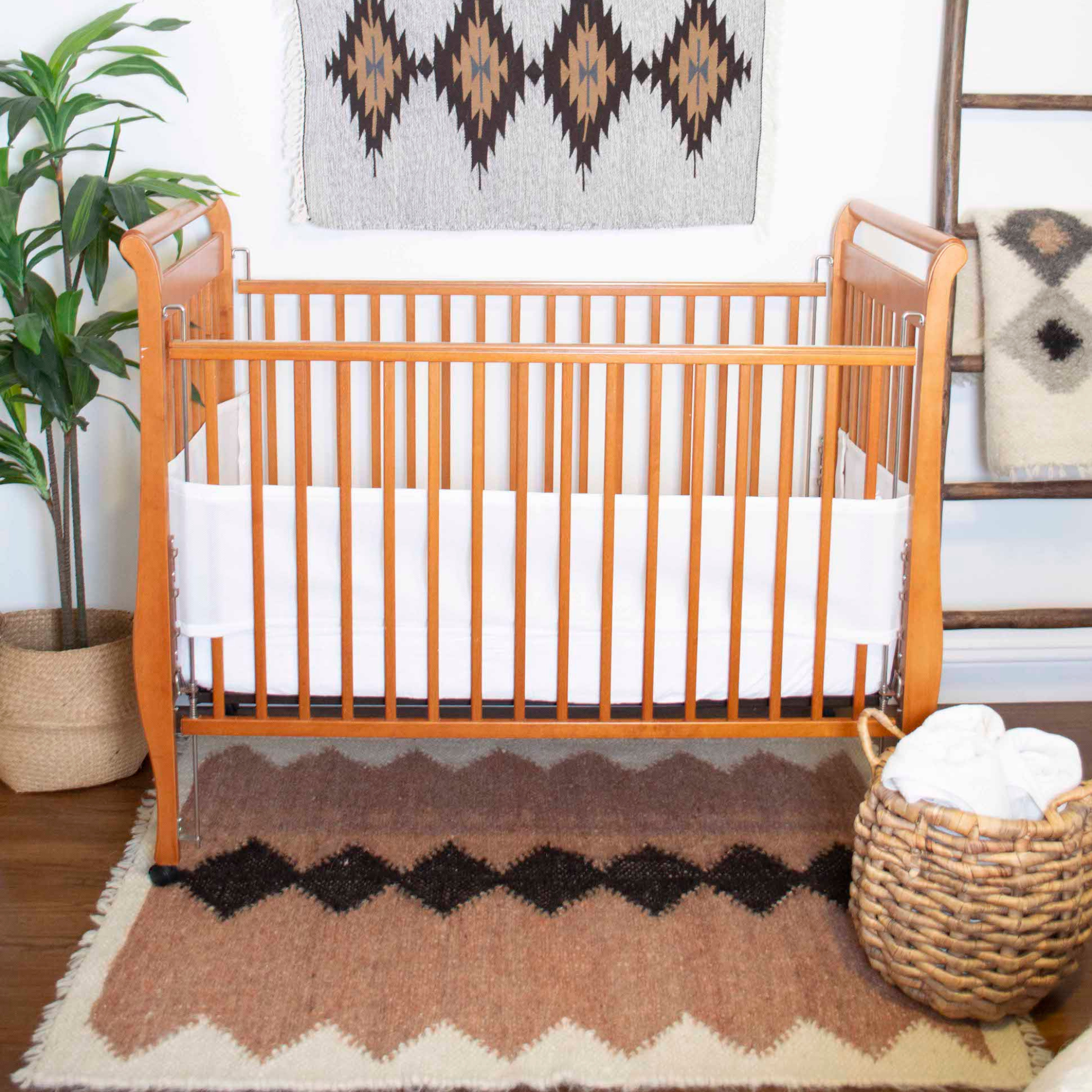 A wooden crib with a white bumper and the Druh (Friend) Wool Statement Throw/Baby Blanket sits by a plant and basket on a patterned rug in a cozy room.