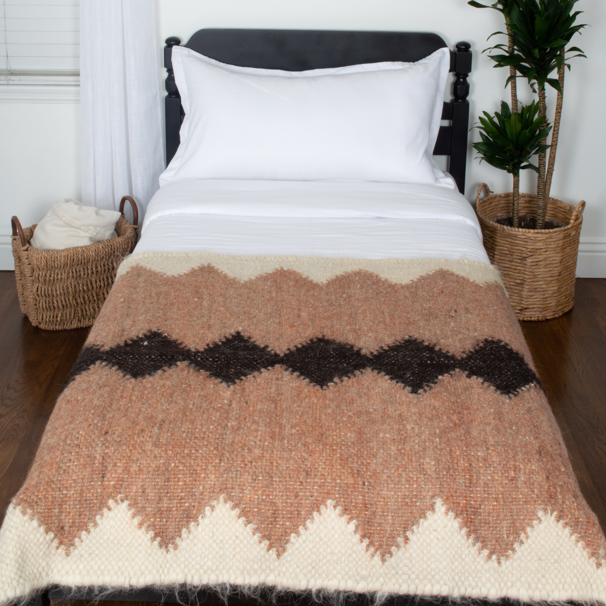 A bed with white linens features the Druh (Friend) Wool Statement Throw as a decorative accent, flanked by woven baskets and a potted plant.