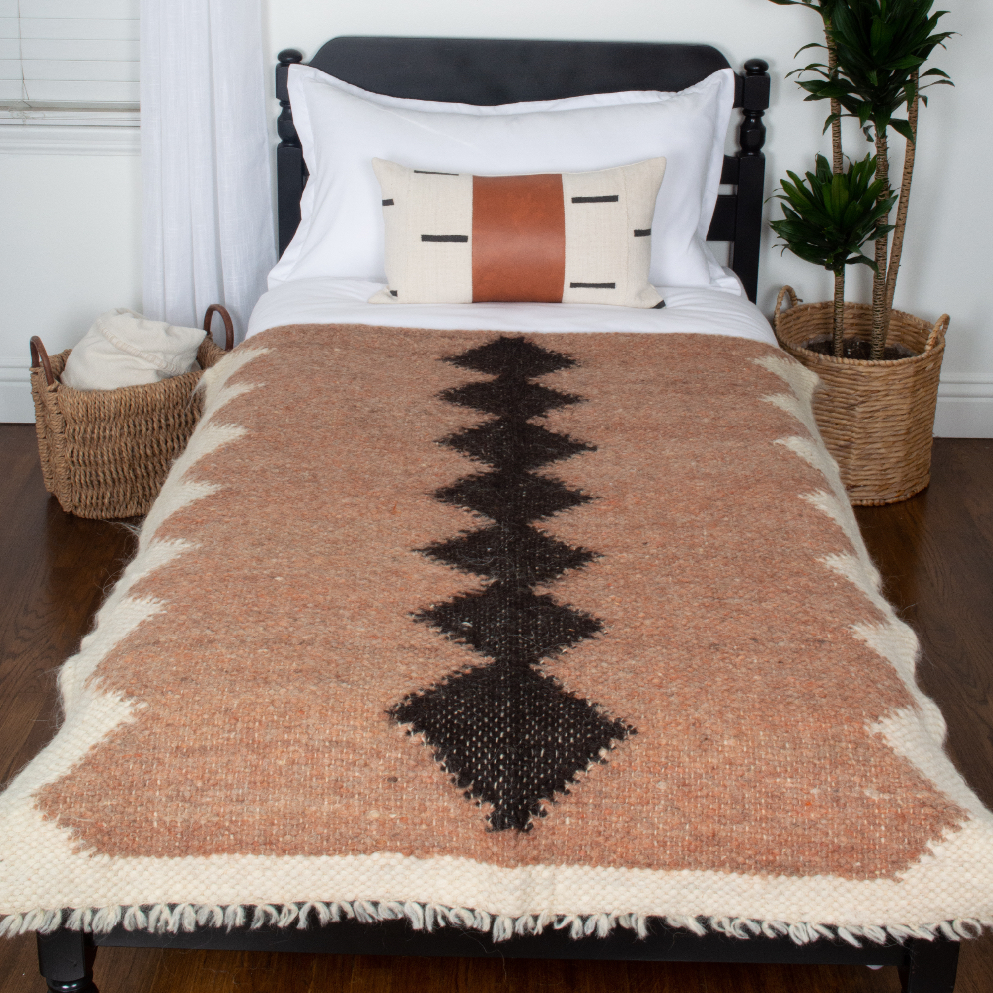 A wooden floor with baskets, a bed featuring a patterned blanket, decorative pillow, and the Druh (Friend) Wool Statement Throw / Baby Blanket, plus a nearby plant.