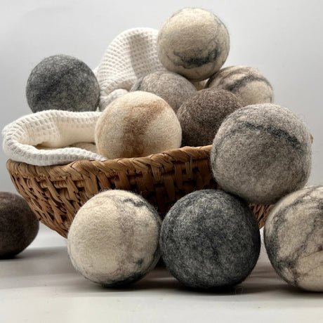 A woven basket and white towel display Fluff Ewes "Bag of Marbles" Premium Dryer Balls—6 dye-free, 100% natural marbleized sheep’s wool balls in gray, white, and brown tones—an eco-friendly laundry essential.