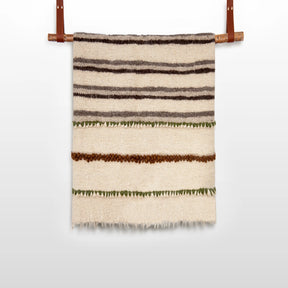 The Faino (Fine) Wool Statement Throw / Baby Blanket is a handwoven cream blanket with brown and green horizontal stripes, displayed on a wooden rod with leather straps, offering a charming and hypoallergenic option for babies.