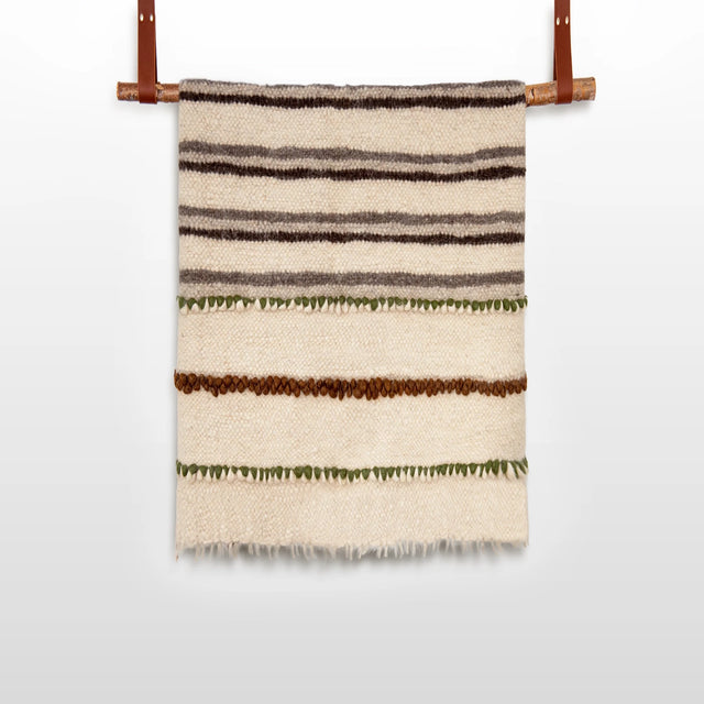 The Faino (Fine) Wool Statement Throw / Baby Blanket is a handwoven cream blanket with brown and green horizontal stripes, displayed on a wooden rod with leather straps, offering a charming and hypoallergenic option for babies.