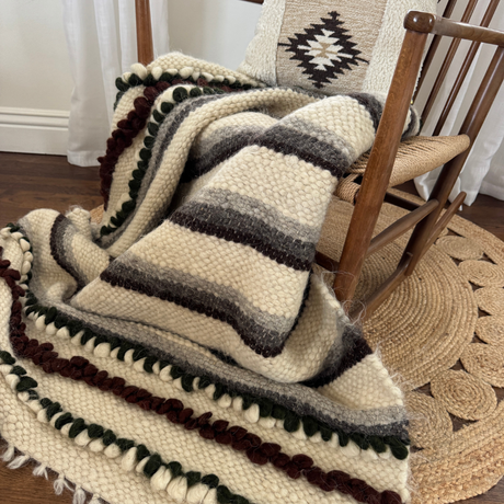 The Faino (Fine) Wool Statement Throw / Baby Blanket, featuring brown, gray, and green stripes, is draped over a wooden rocking chair with a patterned pillow.