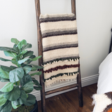 The Faino (Fine) Wool Statement Throw / Baby Blanket hangs on a wooden ladder beside a potted plant and a bed in a cozy room.