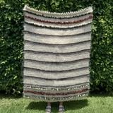 A person holding the Faino (Fine) Wool Statement Throw / Baby Blanket, featuring handcrafted horizontal stripes and textured patterns, stands in front of green bushes.