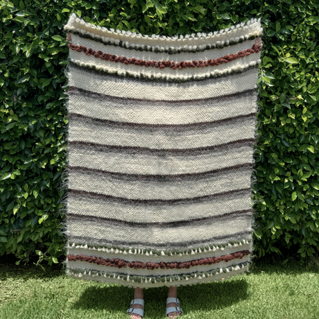 A person holding the Faino (Fine) Wool Statement Throw / Baby Blanket, featuring handcrafted horizontal stripes and textured patterns, stands in front of green bushes.