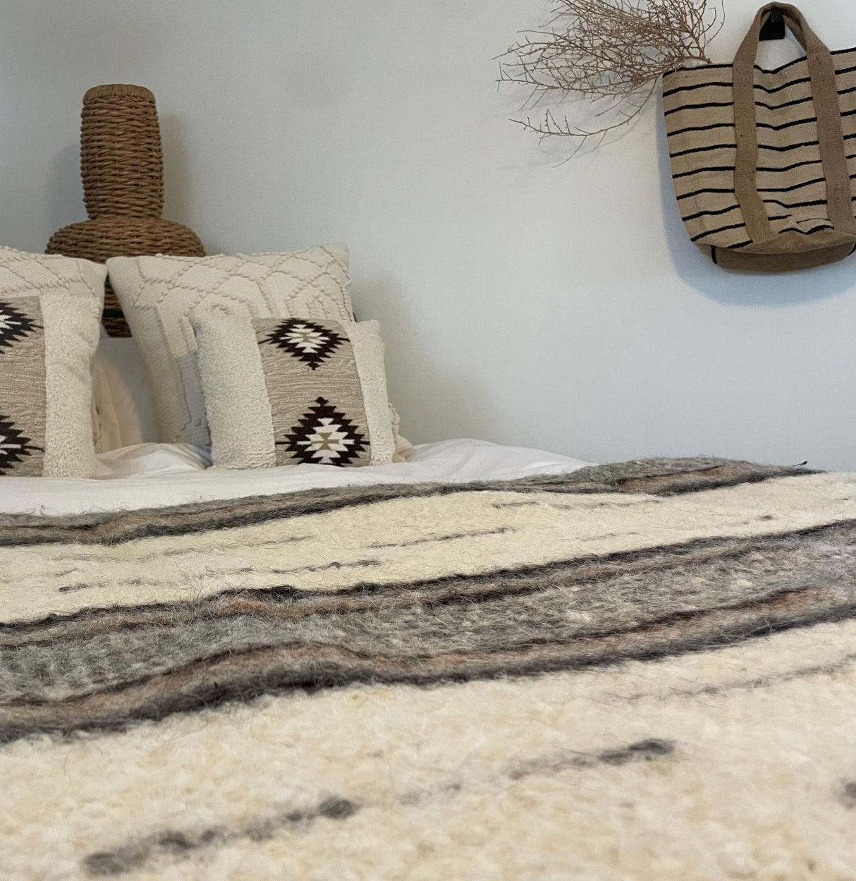 Cozy bed with patterned pillows and the Gratsia (Grace) Wool Statement Bed Blanket; a woven bag and dried branch hang on the wall above.