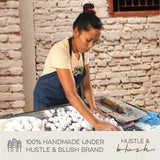 A woman rolls dough by hand on a table covered with Rainbow, which resembles artisan Christmas ornaments, beneath a brick wall.