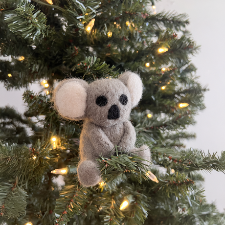 A Hand-Stitched Felted Koala Ornament sits on a lit Christmas tree branch, bringing charming artisan style to your holiday ornament collection.