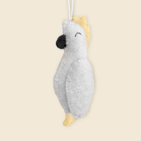The Hand-Stitched Felted Parrot Ornament features a white cockatoo with yellow crest and tail, hanging from a white string. Expertly crafted from sustainable New Zealand wool, it's perfect for fans of artisan Christmas decor.