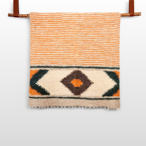The Handmade Organic 100% Sheep Wool Woven Statement Blanket features orange stripes and a black, white, and orange diamond pattern, shown hanging on a rod.