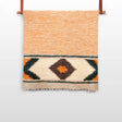 The Handmade Organic Natural 100% Sheep Wool Woven Statement Blanket features orange stripes and a geometric diamond pattern in brown, green, and orange, displayed hanging on a rod.